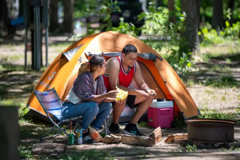 Two people sitting by an unlit fire pit at a campsite in a wooded area. They are next to an orange tent and holding food in their hands. One person is on a folding chair, and there's a cooler behind them. They seem to be enjoying a relaxed conversation.