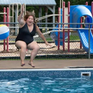 A person in a black swimsuit is captured mid-air while jumping into a swimming pool. A colorful beach ball is also in mid-air next to them. In the background, there is a playground with slides and climbing equipment enclosed by a chain-link fence.