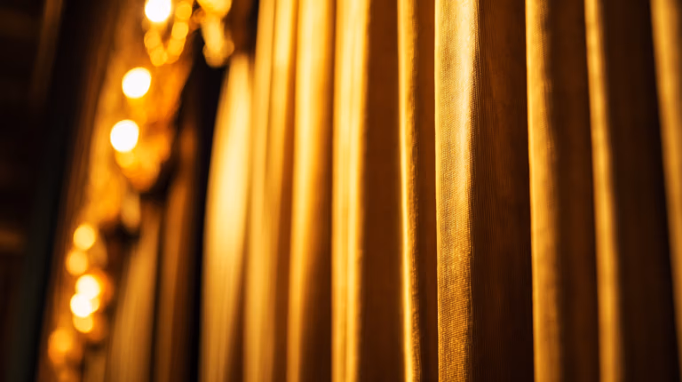 Close-up of golden theater curtains with soft focus. Bright stage lights illuminate the curtain's fabric, casting a warm, inviting glow.