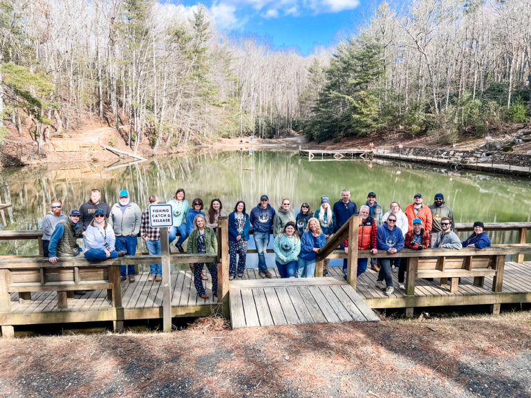 A group of people stands and sits on a wooden structure by a serene, wooded lake. They are dressed in casual clothing and are smiling at the camera. The background features bare trees and a partly cloudy sky. A sign warns of alligators.