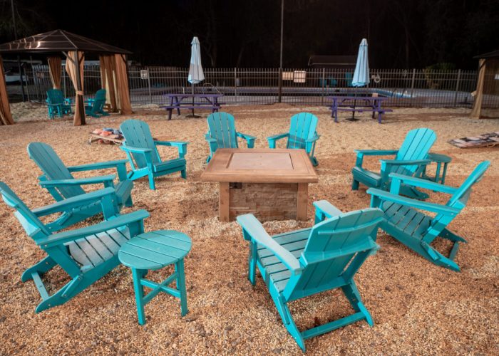 A group of turquoise Adirondack chairs arranged in a circle around a fire pit on a gravel surface, with gazebos, picnic tables, and umbrellas in the background at night.