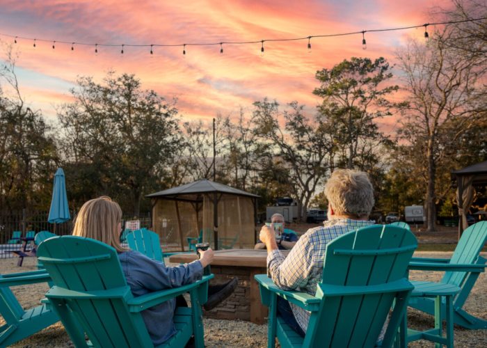 Two people sit in turquoise Adirondack chairs around a firepit, enjoying drinks at sunset. String lights hang above, and trees with sparse leaves are in the background, creating a relaxed outdoor atmosphere.