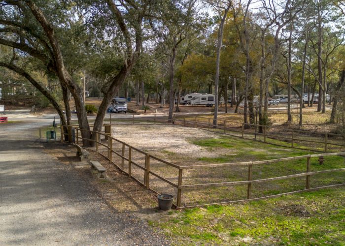 A fenced, gravel RV campsite sits among tall trees in a wooded area, with several RVs parked in the background. The ground is partly grassy, and the atmosphere appears peaceful and quiet.