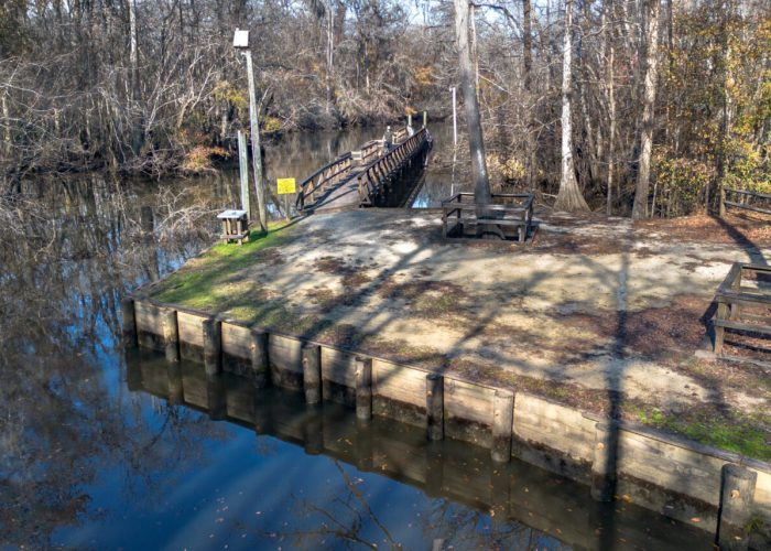 A wooden dock and picnic area sit beside a calm river, surrounded by bare trees in winter. A wooden bridge stretches over the water, and benches and tables are set up on the sandy ground.