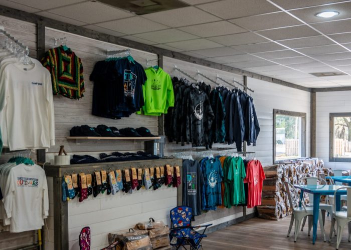 A gift shop with racks of colorful t-shirts, sweatshirts, hats, and bandanas on the left wall. On the right, there are tables and chairs by windows letting in natural light. The shop has wooden floors and a cozy atmosphere.
