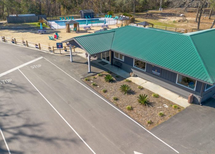 Aerial view of a building with a green roof next to a paved road with "STOP" markings, a small landscaped area, and a playground with a pool, swings, and slides in a fenced area behind the building. Trees surround the area.