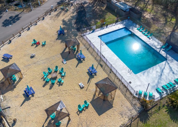 Aerial view of a fenced outdoor swimming pool surrounded by lounge chairs and shaded cabanas on sandy ground, with several seating areas and a nearby road.